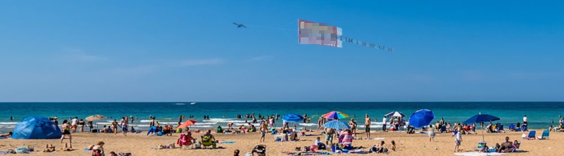 Over a crowded beach, a plane flies a banner ad
